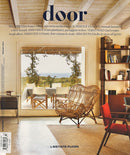 Door Magazine cover featuring a warm modern interior with woven lounge chairs bookshelves and a wide view of trees and ocean through open glass doors